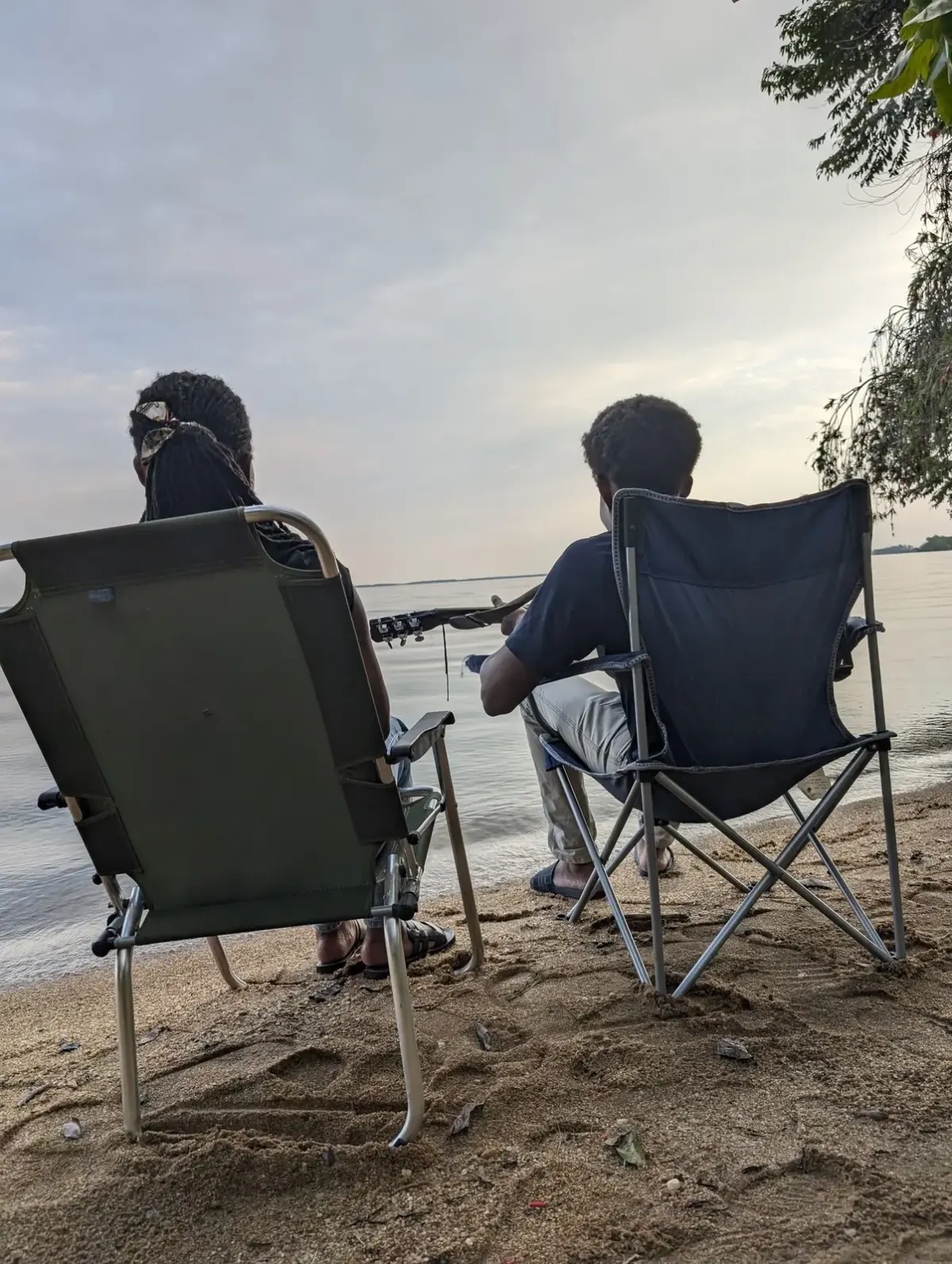 Two guests relaxing in camp chairs on the sandy lakeshore, one playing a guitar