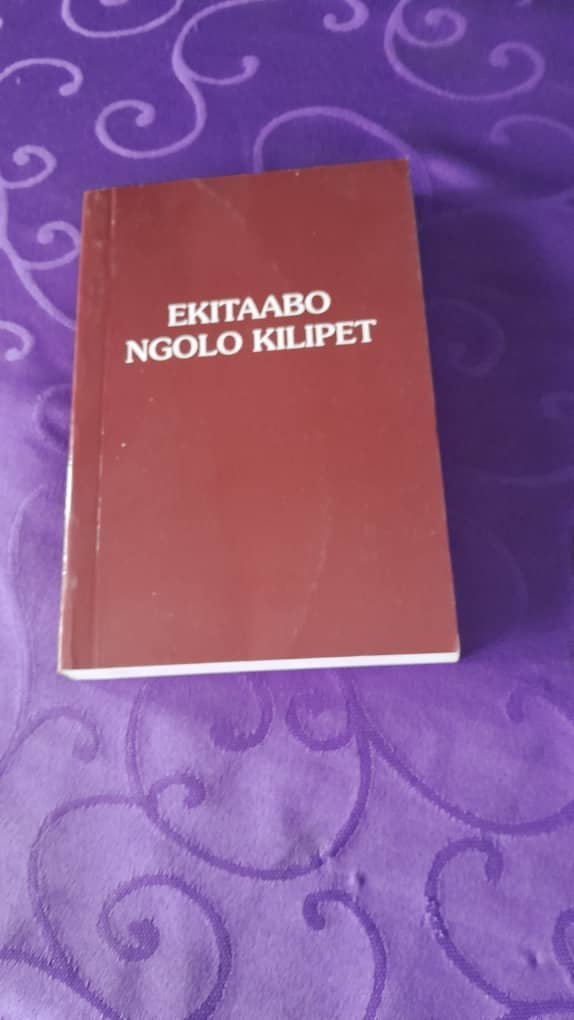 Maroon prayer book titled Ekitaabo Ngolo Kilipet resting on a purple cloth